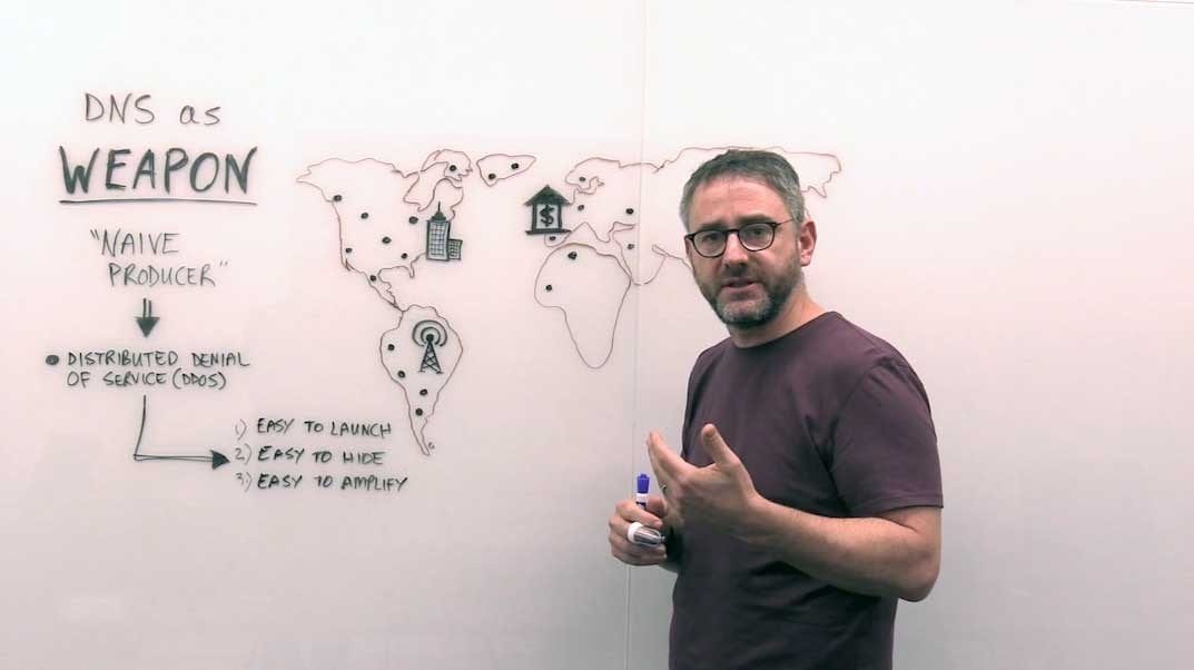 Presenter at whiteboard explaining “DNS as weapon” with DDoS attack diagram and distributed denial of service notes