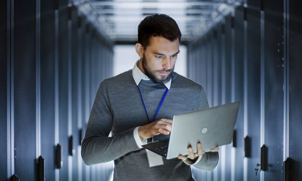Standing man looks at laptop in server room