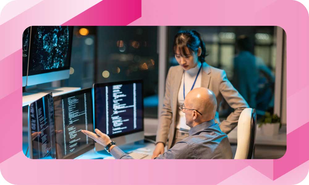 Two network engineers review complex DNS configuration code on multiple monitors in a dim operations room